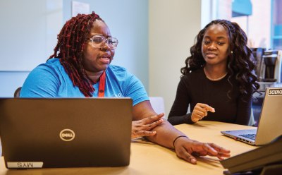 Two individuals engaged in a discussion while working on laptops at a table, background includes an office or co-working space with light blue walls and windows Two individuals engaged in a discussion while working on laptops at a table, background includes an office or co-working space with light blue walls and windows