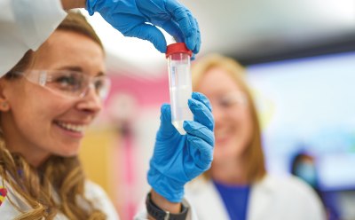 Unlocking Drug Discovery Success for Emerging Biotech Startups A person in blue gloves holding a test tube filled with a clear liquid, another person nearby in a laboratory setting. Background includes various laboratory equipment and bright lighting