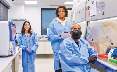 Three lab-coated individuals collaborating in a well-organized laboratory, background includes white cabinets, stocked shelves, and informational posters Three lab-coated individuals collaborating in a well-organized laboratory, background includes white cabinets, stocked shelves, and informational posters