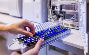 Reference Materials A close-up image of a person's hand inserting a blue and black sample tray into a laboratory instrument. The background shows part of the machine with visible components, emphasizing a laboratory setting.