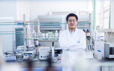 Scientist in pharma lab in a white lab coat stands with arms crossed in a laboratory setting, surrounded by various scientific equipment and glassware.