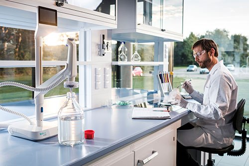 Man in a lab coat sitting at a lab bench with various equipment, including a notebook, pipettes and a Q-POD dispenser filling glassware with water