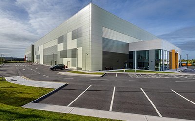 Exterior view of manufacturing facility in Blarney, Ireland, featuring a modern design with large windows and a landscaped parking area, set against a clear blue sky