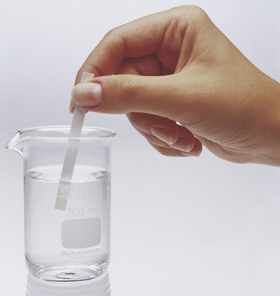 MQuant® test strip in beaker A hand dipping a test strip into a clear glass beaker filled with water. The beaker is marked with measurements indicating 100 ml and is labeled "Made in Germany." The action suggests a chemical or quality testing process.
