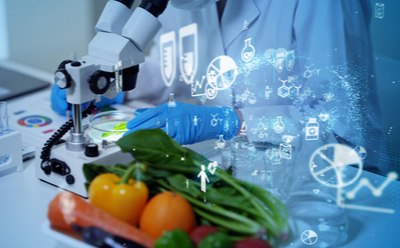 Vitamins A laboratory setting with a focus on food science research. The image shows a robotic arm handling a petri dish, with holographic projections of molecular structures and scientific symbols surrounding it. In the foreground, there is an assortment of fresh vegetables and fruits, including oranges and leafy greens, indicating the study of natural food properties.