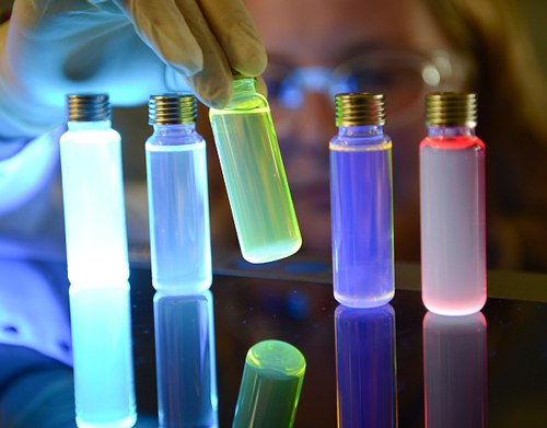 Vials of OLED materials lit up by UV light The image depicts light-emitting materials for organic light emitting diode (OLED).  A person wearing gloves is holding a glowing green vial among other brightly colored glowing vials on a reflective surface. The vials emit various colors, including pale blue, yellowish-green, deep purple, and bright red. The person’s face and protective goggles are subtly outlined by the glow, and each vial has a metallic cap. The reflection of the glowing vials is visible on the surface.
