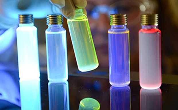 Vials of OLED materials lit up by UV light A close-up image of several glass vials with metallic caps, illuminated by colorful lights. The vials contain liquids that glow in shades of blue, purple, and red, creating a visually striking effect against a reflective surface. A gloved hand is positioned near the vials, suggesting a scientific or experimental context.
