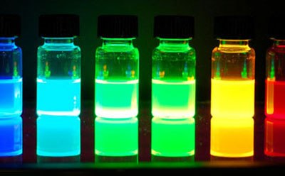 Photoluminescence of alloyed quantum dots Five glass vials filled with brightly colored liquids, displaying shades of blue, green, yellow, and red, illuminated against a dark background.