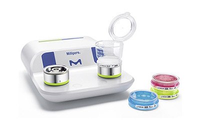 Milliflex Oasis® filtration system for bioburden testing a Millipore microbial filtration system with two filtration units on top and three petri dishes with cultured samples in front. The system is white and blue with the Millipore logo. The petri dishes contain colored media: one blue, one pink, and one yellow.