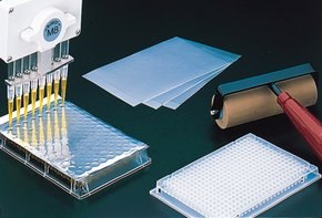 AlumaSeal 96 ® foil A multi-channel pipette dispensing liquid into a well plate, with AlumaSeal 96 ® foil, a stack of filter papers, and a roller bottle nearby, all placed on a dark surface against a black background.