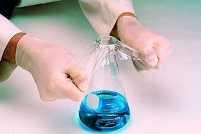 A person wearing lab gloves is applying a sealing film to a flask containing a blue liquid A person wearing lab gloves is applying a sealing film to a flask containing a blue liquid
