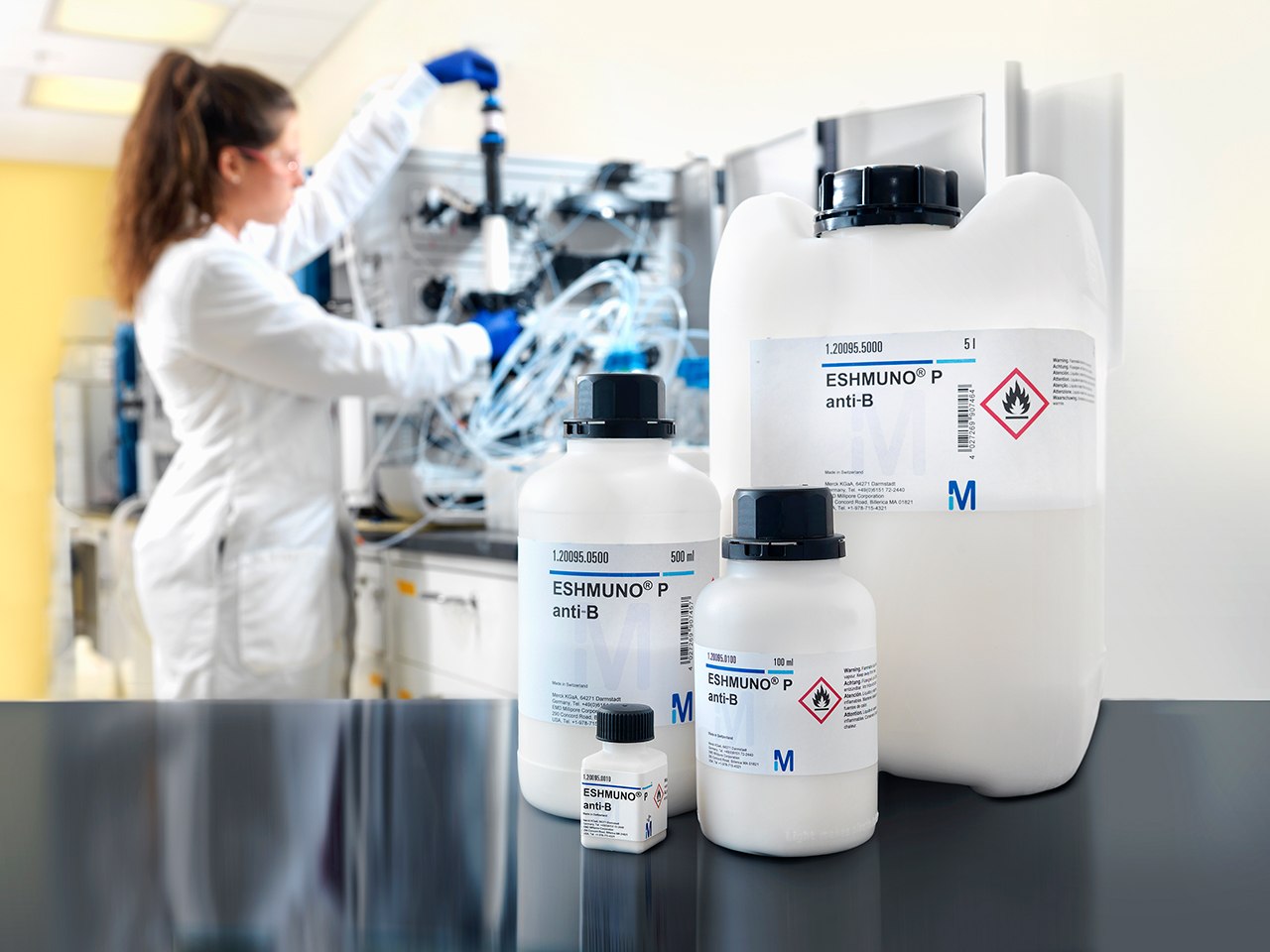 Affinity chromatography with Eshmuno® resins A laboratory setting showcasing the tools and chemicals used in affinity chromatography. In the foreground, three bottles labeled with hazard symbols indicate the presence of flammable and corrosive substances, essential for selective biochemical interactions and purification processes. In the background, a scientist in a lab coat is engaged with complex equipment, likely related to large-scale manufacturing and process development in affinity chromatography.
