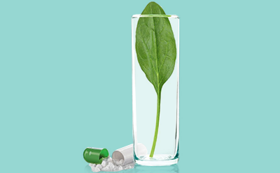 Folate | Mobius® fluid transport and storage systems The image depicts a clear glass containing a large green leaf, surrounded by scattered pills and capsules against a teal background. One of the capsules is open, revealing its powdered contents.