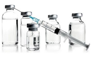 Bioprocessing & Formulation Raw Materials A medical syringe with a needle, partially filled liquid, is positioned diagonally in front of five vials of various sizes with metallic caps against a white background.