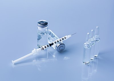 A sterile setting showcasing pharmaceutical preparations for injectables and parenteral formulations. The following elements are visible: 1) A clear vial with a sealed cap, containing a liquid solution. 2) A white syringe with measurement markings is positioned next to the vial. 3) Three clear ampoules stand upright on the reflective surface. The entire setup is placed on a reflective surface.