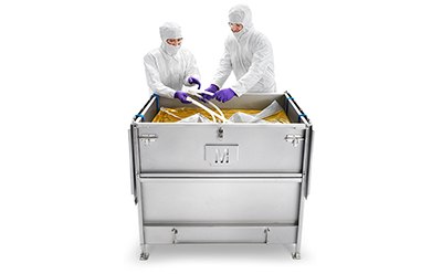 Two individuals in protective white suits, gloves, and head covers are standing at a stainless steel cart with large gold-colored bags inside it. Two individuals in protective white suits, gloves, and head covers are standing at a stainless steel cart with large gold-colored bags inside it.
