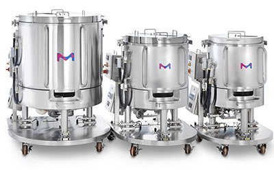 Three stainless steel bioreactor vessels of varying sizes on wheeled stands, each with a blue and pink ‘M’ logo on the front. Three stainless steel bioreactor vessels of varying sizes on wheeled stands, each with a blue and pink ‘M’ logo on the front.