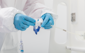 Connect the NovaSeptum® SURe sampling assembly via an AseptiQuik® connector A person in protective gear, including gloves, handling a medical syringe and vial. The individual is dressed in white, with blue gloves, and is focused on the medical equipment.