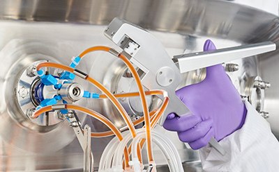 A person in a lab coat and purple gloves, who appears to be adjusting equipment with various tubes connected to it. The equipment includes multiple orange and blue tubes, indicating a fluid transfer or analysis apparatus. A person in a lab coat and purple gloves, who appears to be adjusting equipment with various tubes connected to it. The equipment includes multiple orange and blue tubes, indicating a fluid transfer or analysis apparatus.