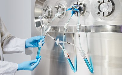 A person in a lab coat and blue gloves handling clear tubing filled with a bright blue liquid, connected to a metallic circular machine with various knobs. The person is conducting an experiment or process involving the transfer of the blue liquid. A person in a lab coat and blue gloves handling clear tubing filled with a bright blue liquid, connected to a metallic circular machine with various knobs. The person is conducting an experiment or process involving the transfer of the blue liquid.