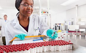 Scientist looking at a vial in her hand. There is a collection of vials in front of her. In the background another scientist walks towards her. 