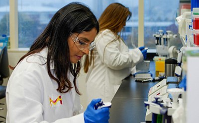 A scientist sits at the lab bench wearing lab goggles, a lab coat, and gloves. She is pipetting. Behind her another scientist wearing a lab coat and goggles is using a multichannel pipettor. There are instruments and pipette tips on the right of the image. A scientist sits at the lab bench wearing lab goggles, a lab coat, and gloves. She is pipetting. Behind her another scientist wearing a lab coat and goggles is using a multichannel pipettor. There are instruments and pipette tips on the right of the image.