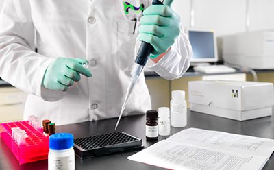 A laboratory technician in a white lab coat and green gloves is using a pipette to transfer liquid into a black microplate. Various laboratory supplies, including bottles of reagents and a pink rack for test tubes, are visible on the table, alongside a document with printed instructions. In the background, laboratory equipment is partially visible. A laboratory technician in a white lab coat and green gloves is using a pipette to transfer liquid into a black microplate. Various laboratory supplies, including bottles of reagents and a pink rack for test tubes, are visible on the table, alongside a document with printed instructions. In the background, laboratory equipment is partially visible.