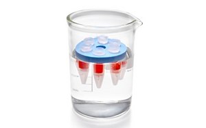 D-Tube™ Dialyzers A clear laboratory beaker filled with colorless liquid, containing a small cylindrical container with a red liquid inside. The container is topped with a blue lid, floating on the surface of the liquid in the beaker. The beaker has measurement markings on the side, indicating its volume.