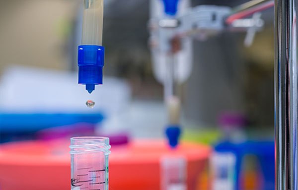 Protein purification A close-up of a laboratory setting. The main focus is on a blue pipette dispensing a droplet into a small glass container partially filled with a pink-colored liquid. The background, although blurred, suggests the presence of various other laboratory instruments or containers.