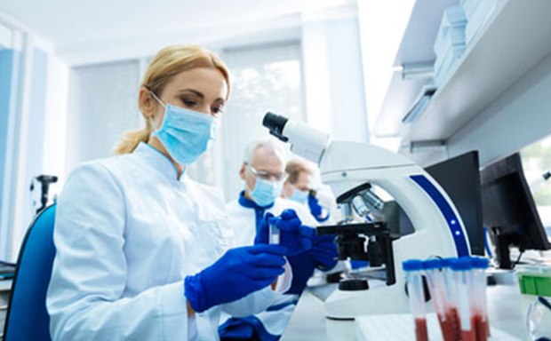 Structural Genomics Consortium (SGC) An individual working in a well-lit laboratory setting, wearing a white lab coat and blue gloves, indicating a sterile or clean environment. Individual using a modern microscope, focusing on something with their hands adjusting the knobs. Test tubes filled with red liquid are visible in the foreground, suggesting some biological or chemical testing.