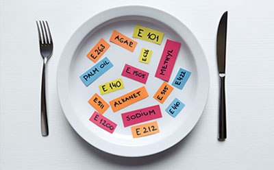 E number standards for food and beverage testing Title: Food additives encoded with the “E numbers” A place setting including a plate, fork, and knife. The plate features colorful labels with text related to food additives, such as “E120,” “SODIUM,” and “E122.”
