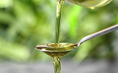 Olive Oil Pouring into Spoon The image appears to show a viscous, golden liquid—likely oil—being poured into a spoon. The blurred green background suggests a natural setting, which could relate to themes of cooking, food ingredients, health supplements, or natural products.