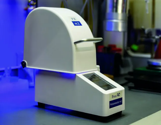 Penn PhD Photoreactor A Penn PhD Photoreactor M2 with a white arched top emitting blue light, placed on a lab bench with industrial equipment in the background.