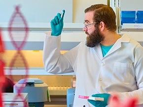 Lab technician in a white coat and teal gloves holds a small tube and pink rack, gesturing in a laboratory with shelves of supplies and red Sigma‑Aldrich boxes in the foreground.
