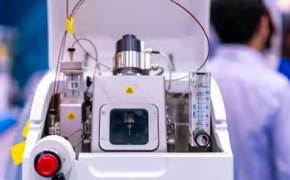 Protein Mass Spectrometry Tools  A mass spectrometer sits in focus in the center of the photo. Behind and to the right stands a scientist with his back turned