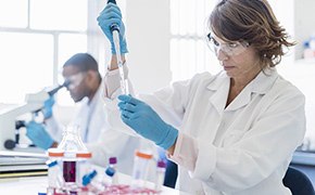 Glycobiology Profiling Tools Female scientist wearing white lab coat, blue nitrile gloves, and safety goggles pipetting a protein solution into a centrifugal tube. Male scientist wearing same protection clothing looking into microscope is visible in the background.