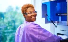 Quality Control Testing Services Female scientist in purple lab coat and safety goggles in molecular lab smiles into camera as she works with instrument preparing biosafety testing samples.