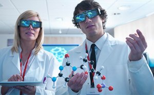 Two scientists wearing 3D glasses Two scientists, a woman and a man, stand side by side in a brightly lit, modern laboratory wearing white lab coats and large black 3D glasses. The woman, with straight blonde hair, holds a tablet in both hands and wears a red lanyard. The man has short brown curly hair and is holding a colorful molecular model made of plastic balls and sticks in black, blue, white, red, and purple, representing different atoms and bonds. He also wears a red lanyard and a name badge over a white shirt and black tie. Behind them, a digital screen displays a bright blue hexagonal molecular structure. The room is well-lit with soft white ceiling lights, pale wood walls, and a partially visible computer station in the background, giving the setting a clean and high-tech atmosphere focused on molecular research or visualization.