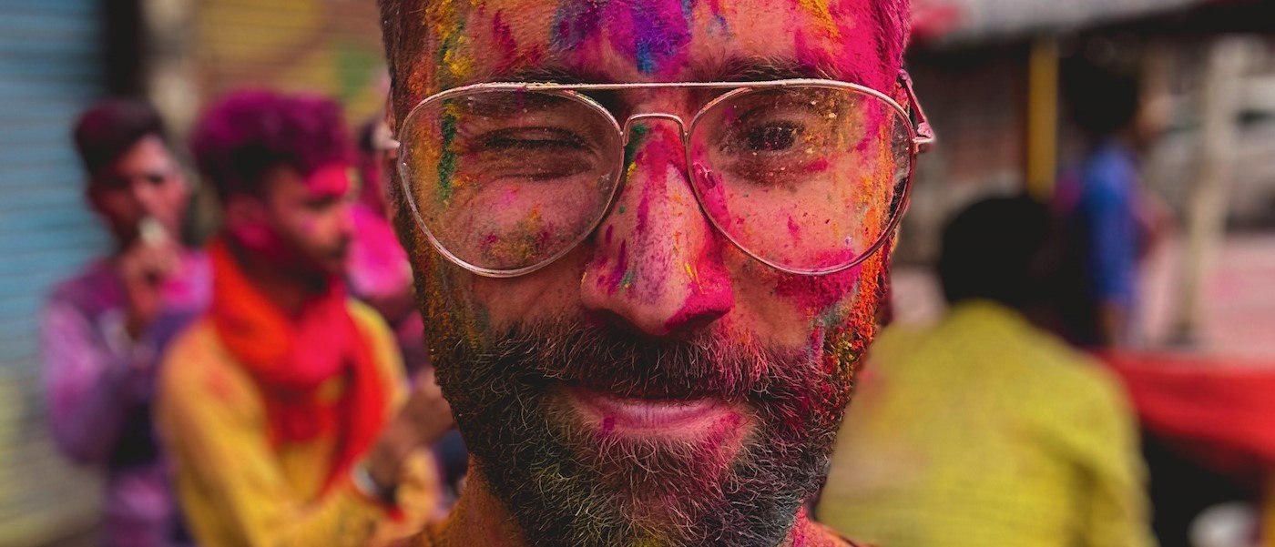 Andre Silva A close-up of André Silva’s face. Silva wears glasses and squints one eye. A number of colors appear on his face, evidence that he participated in a Holi celebration.