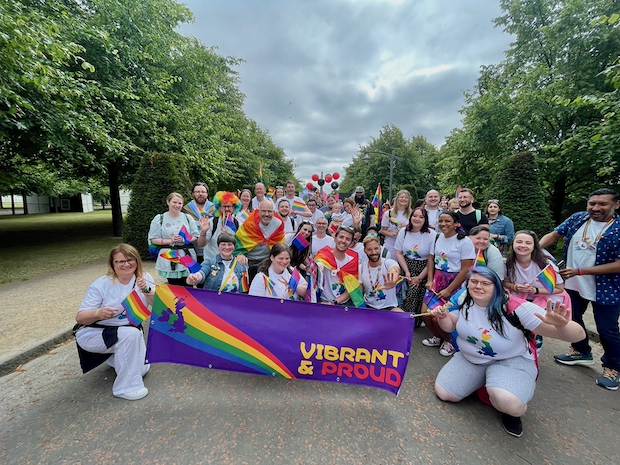 Cork Pride 2022 A group of about 30 employees holds up a banner that reads “Vibrant and Proud” with a rainbow flag on the side. They hold rainbow flags and many are smiling.
