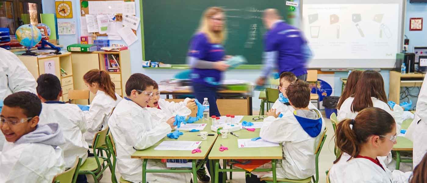Curiosity Lab Classroom Scene A handful of students sit at their desks wearing lab coats and gloves. Two staff volunteers walk around to support students.
