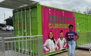 Danielle Chavis Curiosity Cube  Danielle Chavis wears a SPARK t-shirt and stands in front of the Curiosity Cube which is bright green, has images of two young scientists with the words “Breakthroughs Begin with Curiosity” on the front.