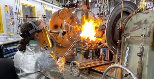Glass Shop Glassblower A round-bottom flask sits in a lathe and a glassblower directs a targeted flame onto the flask’s neck. The glassblower wears protective clothing, including a specialized jacket, gloves and face shield.