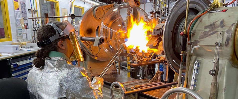 Glassblower works on flask  A round-bottom flask sits in a lathe and a glassblower directs a targeted flame onto the flask’s neck. The glassblower wears protective clothing, including a specialized jacket, gloves and face shield.