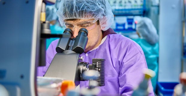 Innovative products expertise - Scientist using microscope Scientist wears a purple lab coat, protective eyewear and a hair net while peering into a microscope.