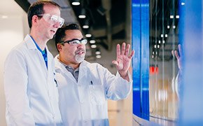 One scientist explaining new innovation to another scientist in front of a blue window One scientist explaining new innovation to another scientist in front of a blue window