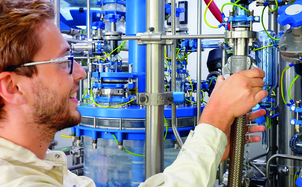 A male production worker in the Schaffhausen pilot plant facilities is connecting a solvent hose to the reactor system, preparing for upcoming manufacturing activities. A male production worker in the Schaffhausen pilot plant facilities is connecting a solvent hose to the reactor system, preparing for upcoming manufacturing activities.