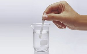 Dipping MQuant® Test Strip A hand is holding a test strip above a beaker filled with clear liquid. The beaker is labeled with a measurement of 100 ml and is placed on a light background. The test strip is being lowered into the water.