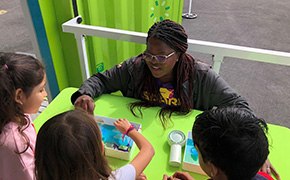 Curiosity Cube  A group of children is engaged in a hands-on science activity at a bright green table, where they are exploring a water filtration experiment. The facilitator, wearing a SPARK™ shirt, demonstrates the process while the children attentively observe and participate, fostering an interactive learning experience outdoors.