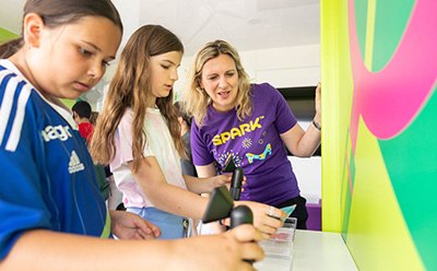 SPARK™  Three children are engaged in an interactive activity at a colorful educational booth, with one child holding a device while another observes intently. An adult, wearing a purple SPARK™ volunteer shirt, is guiding them through the activity, promoting hands-on learning and engagement. The vibrant background features bright colors and patterns, enhancing the lively atmosphere of the event.